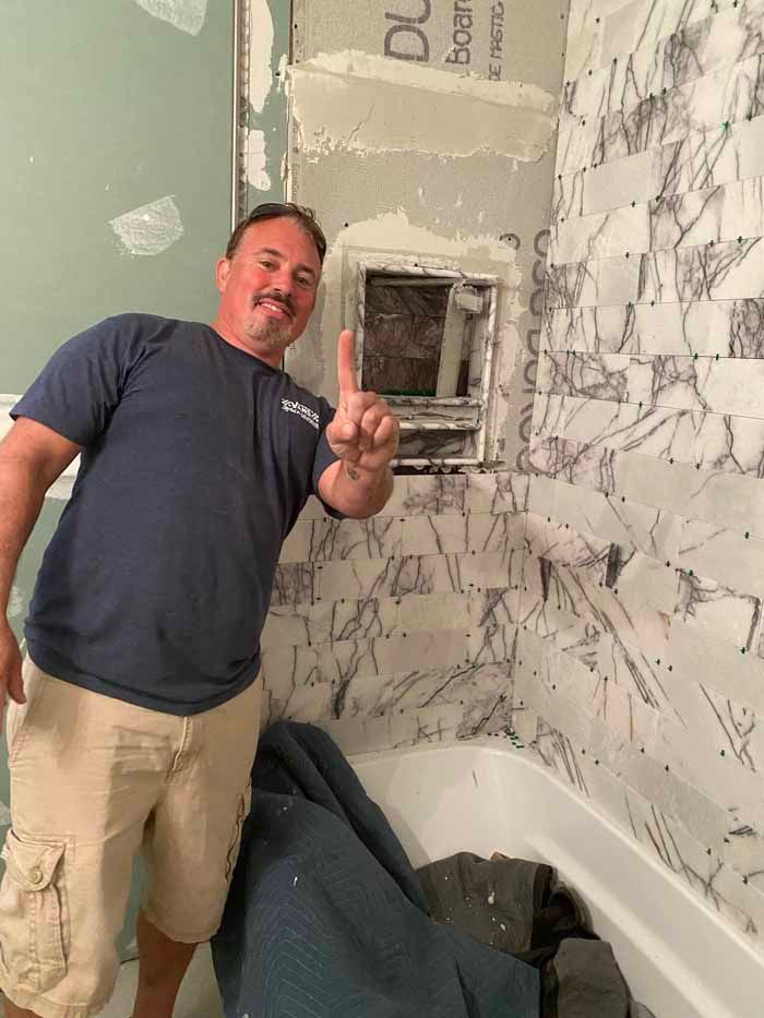 Man smiling and pointing beside a rough wall with a recessed box in a bathroom under renovation
