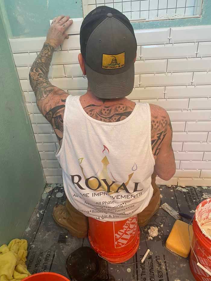 Person painting a tiled wall in a small bathroom, wearing a white tank top and cap.
