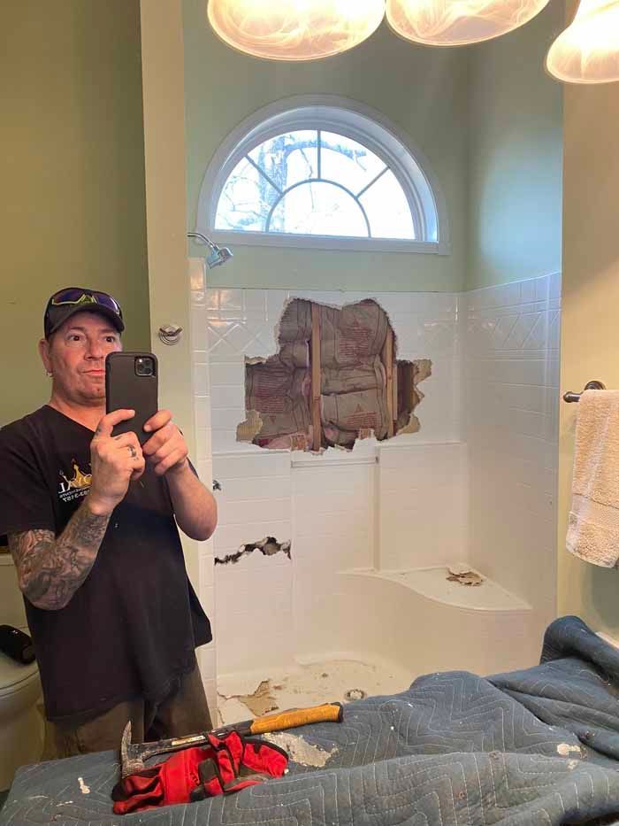 Person taking a mirror selfie in a bathroom with a damaged section of wall and shower area.