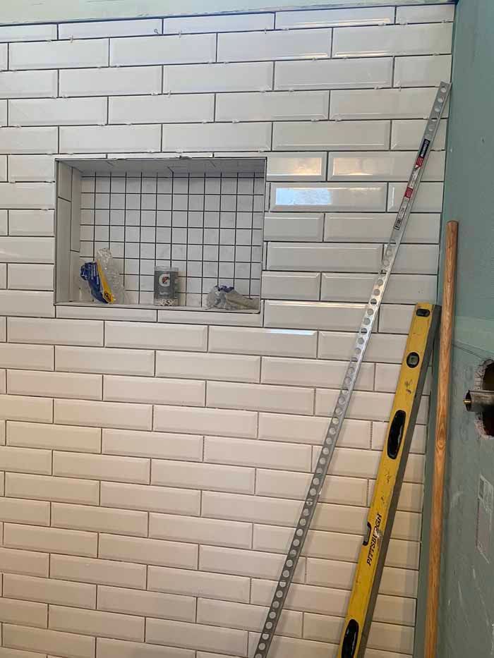 White tiled wall with a small recessed niche, tools inside, and a ladder and level leaning nearby.