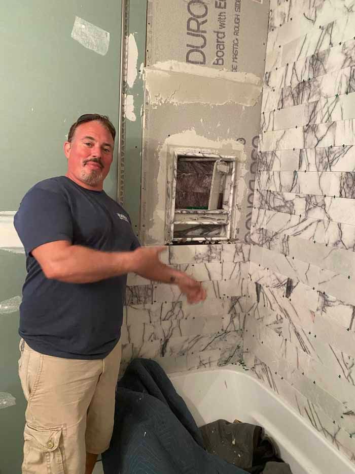 Man standing in a partially tiled bathroom, pointing at a rough wall area beside a bathtub.