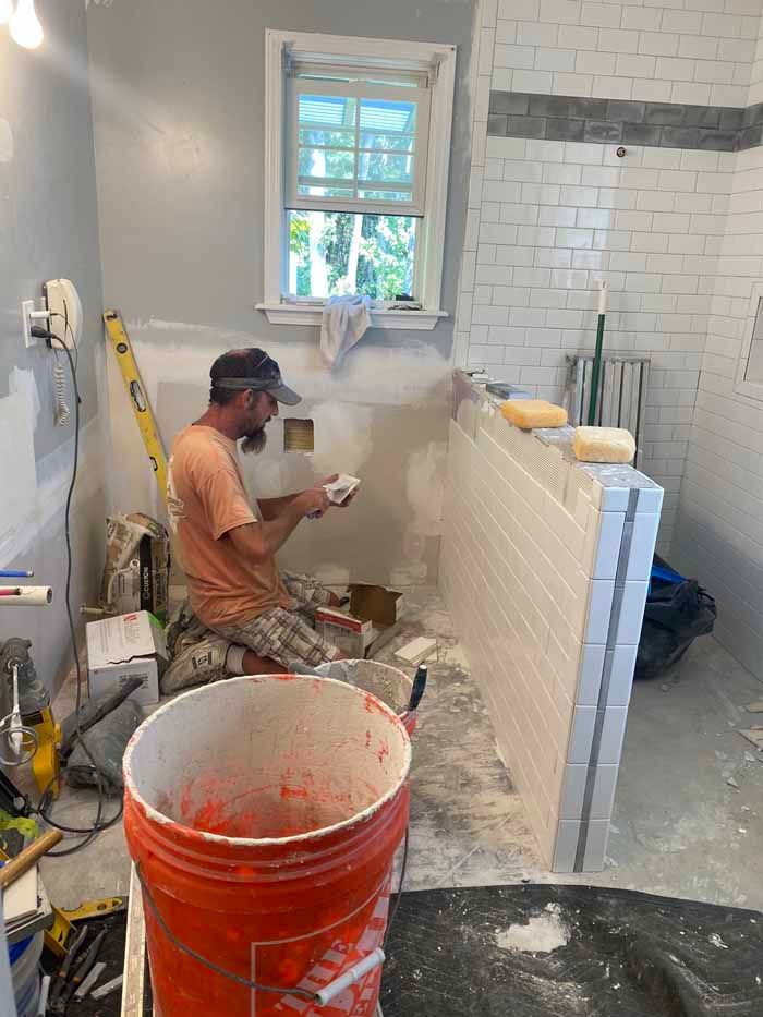 Worker installing white tile in a bathroom, with orange bucket and tools in the foreground.