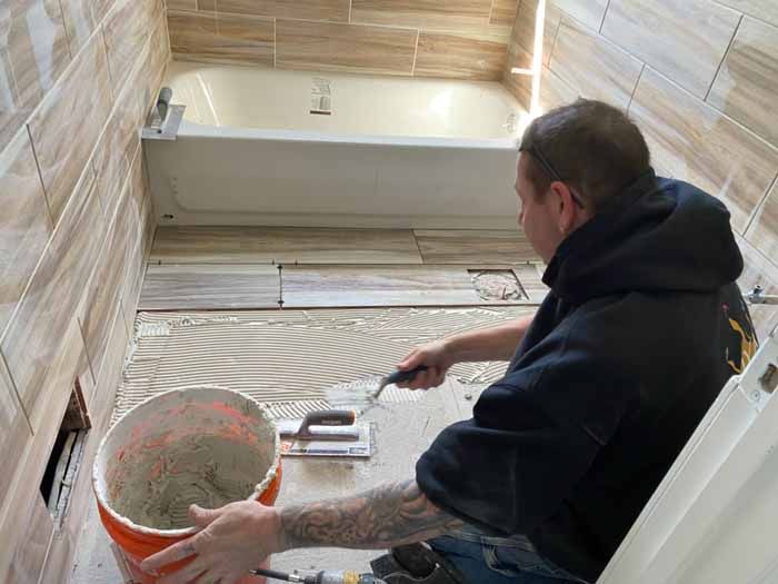 Person troweling mortar on a bathroom floor beside a bathtub and bucket