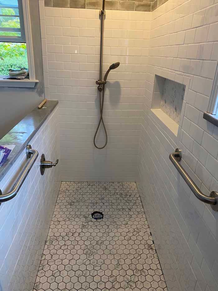 White-tiled walk-in shower with handheld showerhead, bench, grab bars, and pebble floor.
