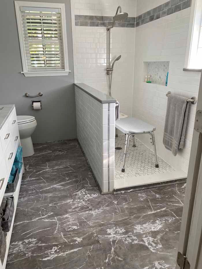Accessible bathroom with white tiled shower, gray marble floor, wall bench, and grab bars.