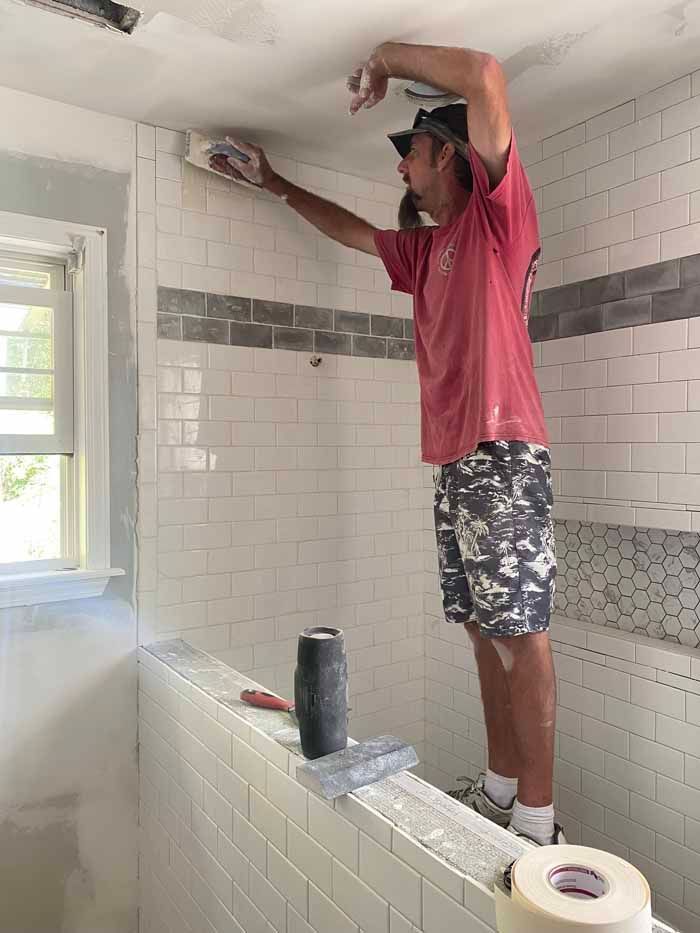 Man tiling a bathroom wall above a tub, reaching up with a trowel in a partially renovated shower area