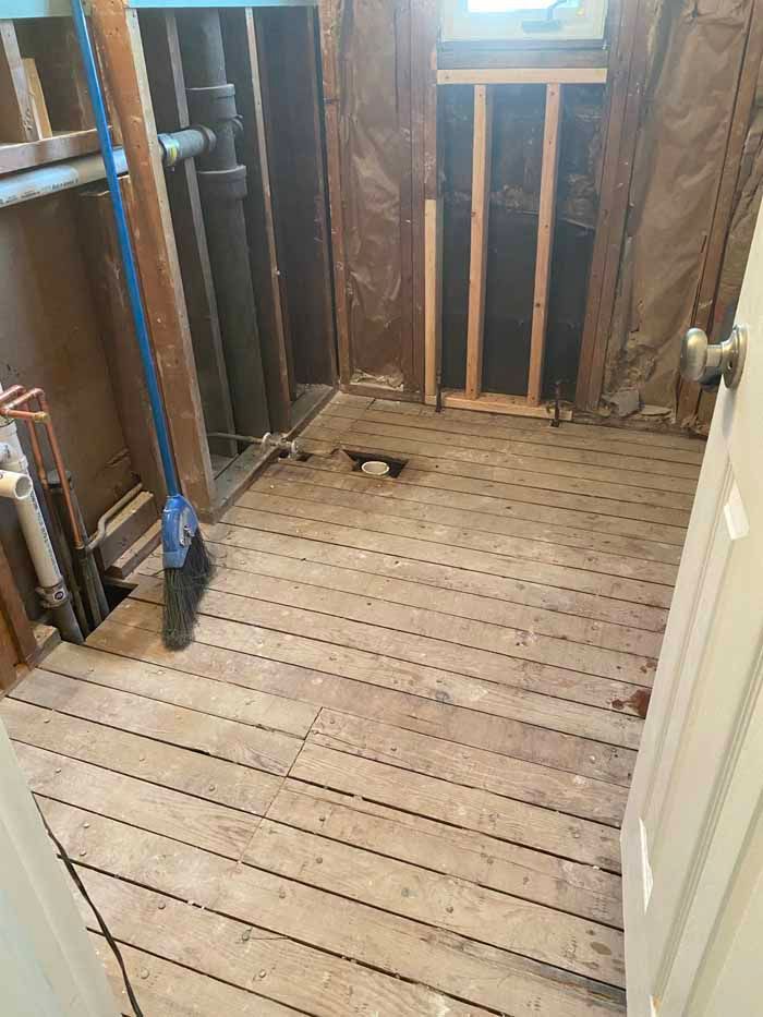 Bare wooden room under renovation with exposed studs, pipes, and an open white door.