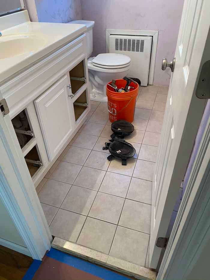 Bathroom floor with a red bucket and black bag beside the toilet and sink cabinet