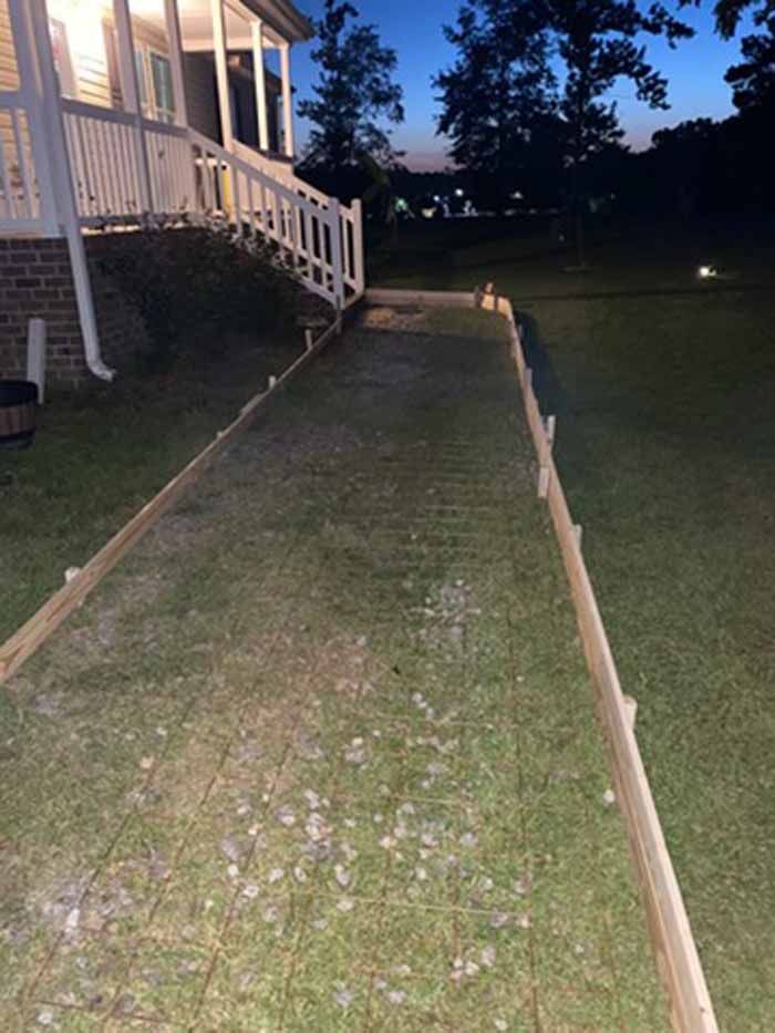 Grassy side yard beside a house at dusk, bordered by wooden edging and lit by a small path light.