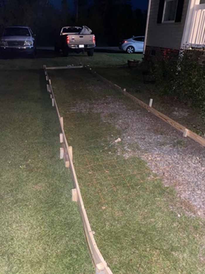 Nighttime front yard with a narrow concrete path bordered by stakes and string, leading to a house drive and parked cars.