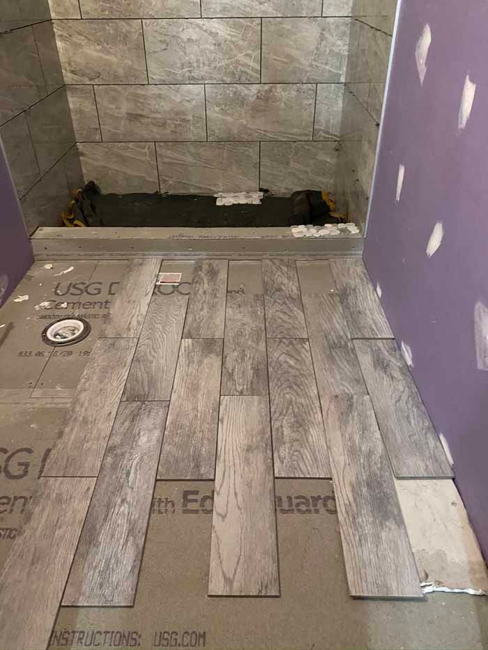 Bathroom shower area under renovation with gray tile walls, new floor tiles, and exposed plumbing.