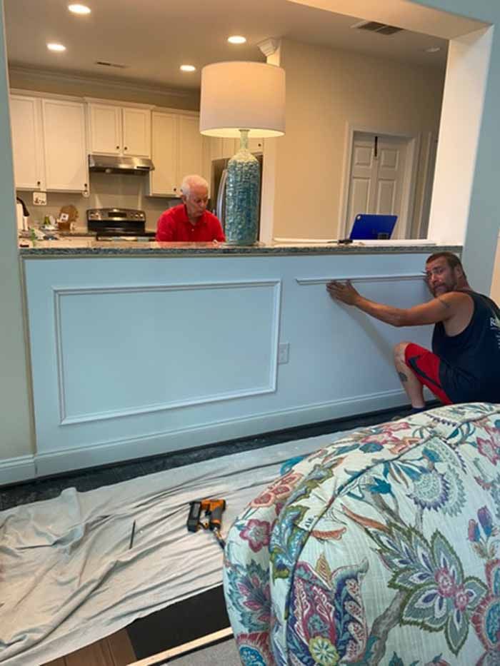 Two men installing a white wall panel in a kitchen passthrough, with tools and a covered floor nearby