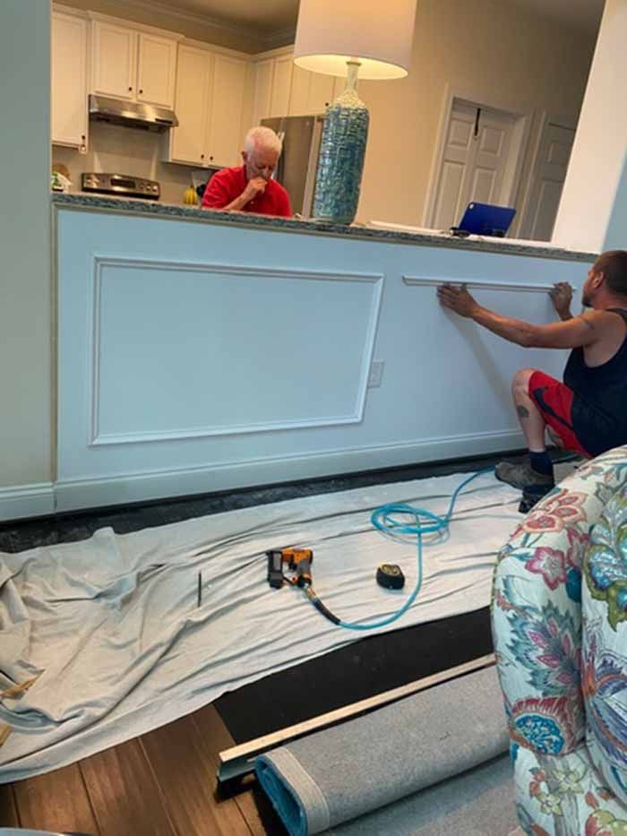 Two people installing a white wall panel in a home interior with tools and drop cloths on the floor