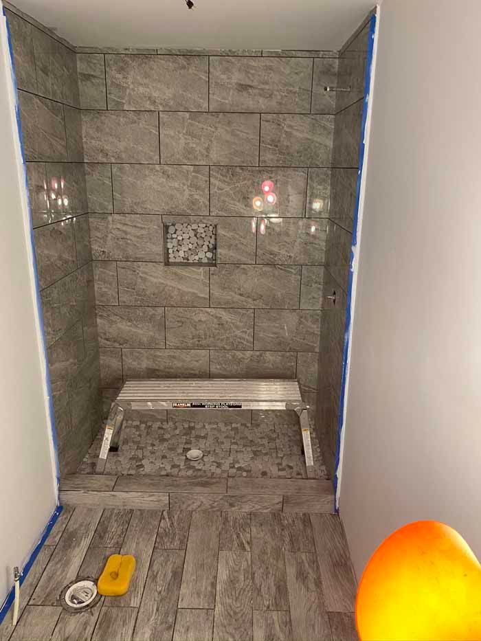 Partially tiled shower stall with gray stone walls and floor, unfinished plumbing, and a drain opening