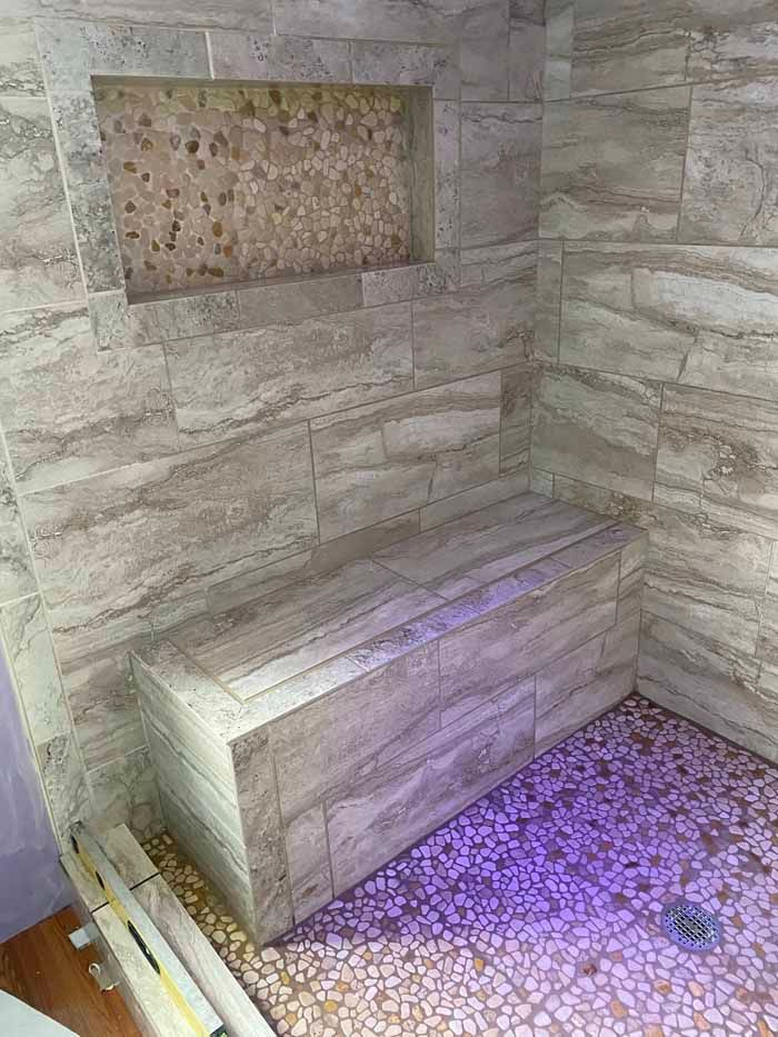 Stone shower with built-in bench and pebble floor, lit by purple light.
