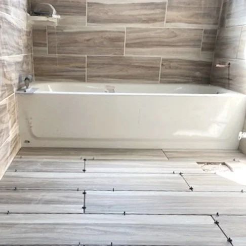 White bathtub in a tiled bathroom with wood-look floor tiles and gray wall tiles.