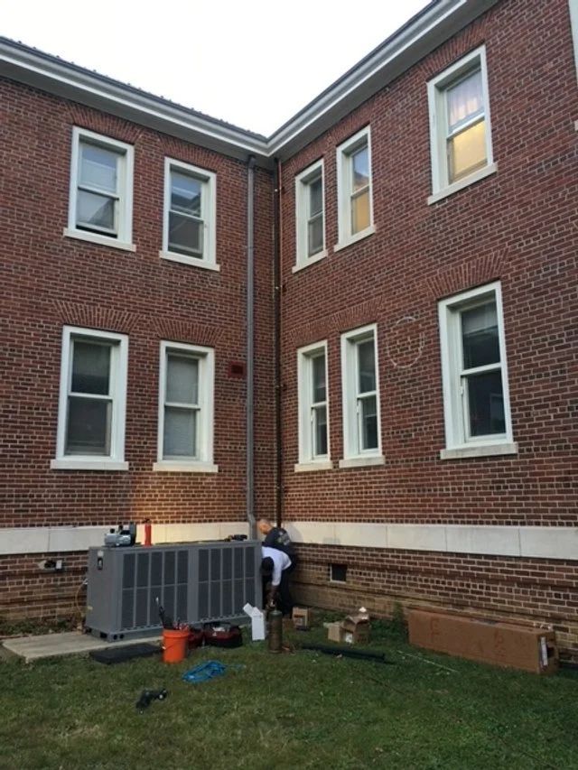 Two people working beside a gray HVAC unit in a brick courtyard between apartment buildings.