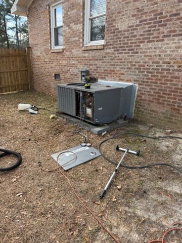 Broken outdoor HVAC unit beside a brick house, with detached panel and tools on the ground.