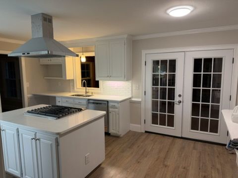 Modern white kitchen with island, stainless hood, light wood floor, and French doors to the right