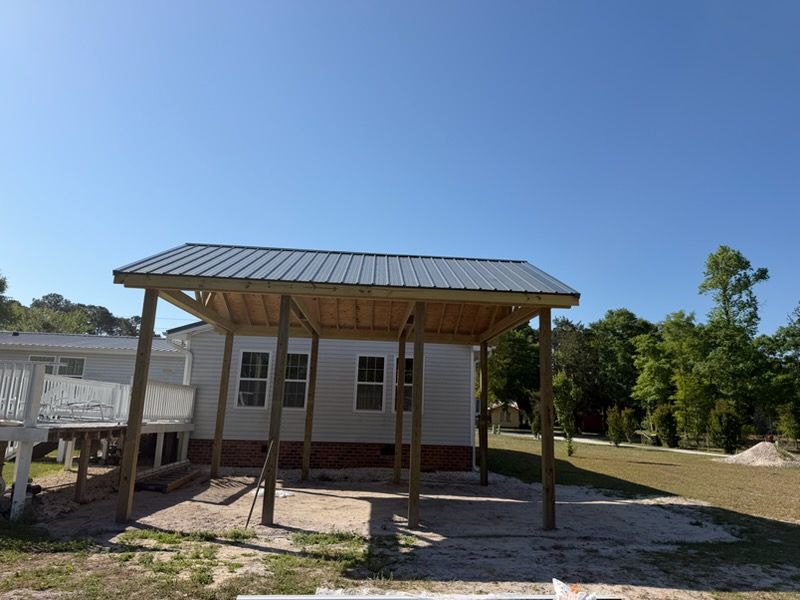 Small white house with a metal roof and front porch on a sunny, open lot surrounded by trees.