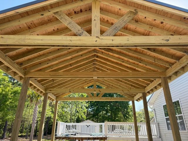 Wooden pergola-style pavilion with exposed beams over a deck, surrounded by trees and a house.
