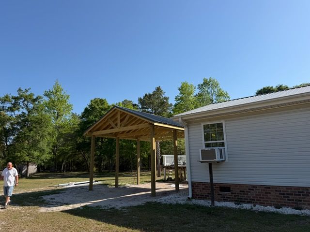 Covered wooden patio beside a white house with an air conditioner, under a blue sky, with a person walking nearby