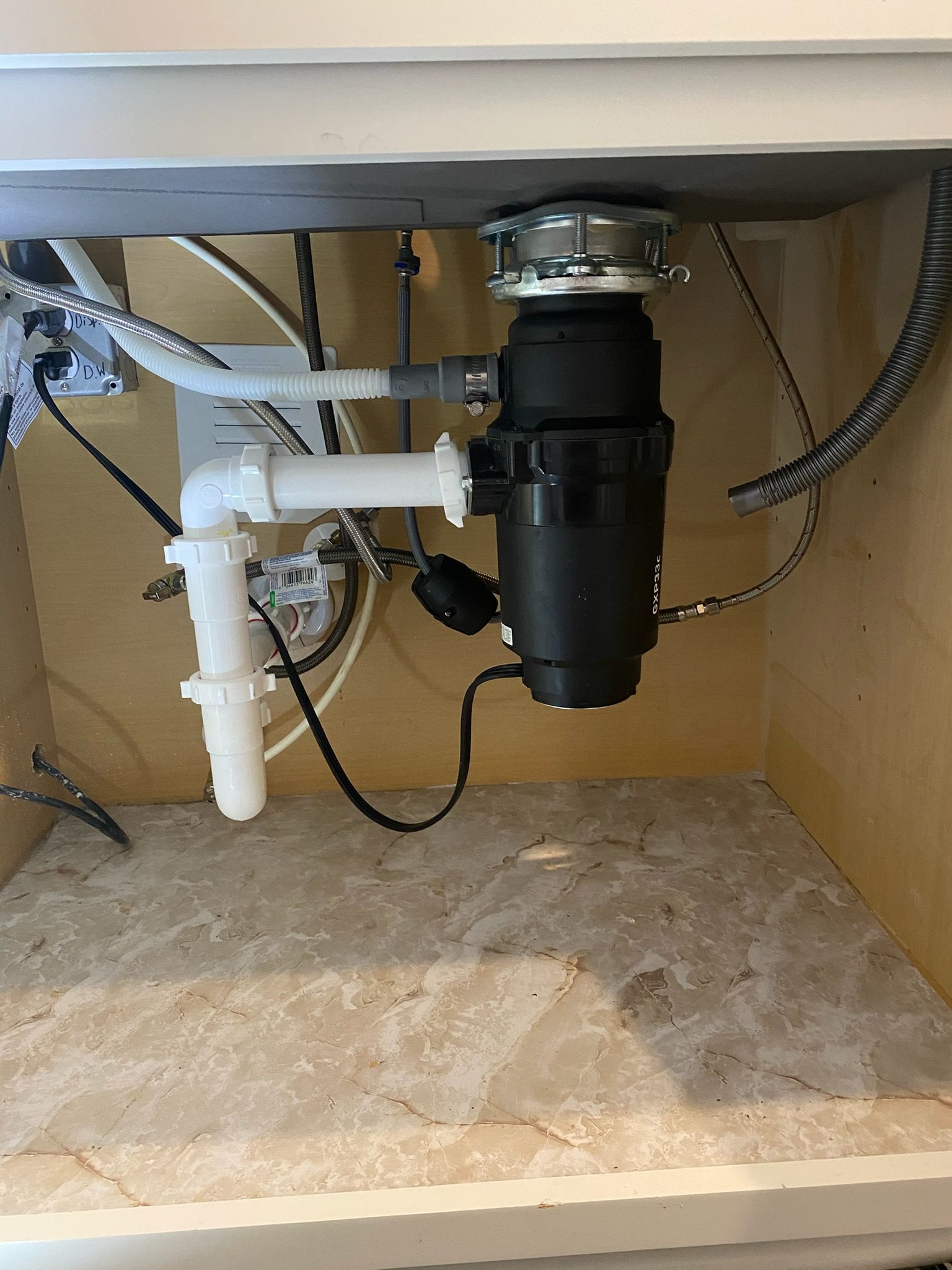 Under-sink plumbing with white PVC pipes and a black garbage disposal against a beige cabinet interior