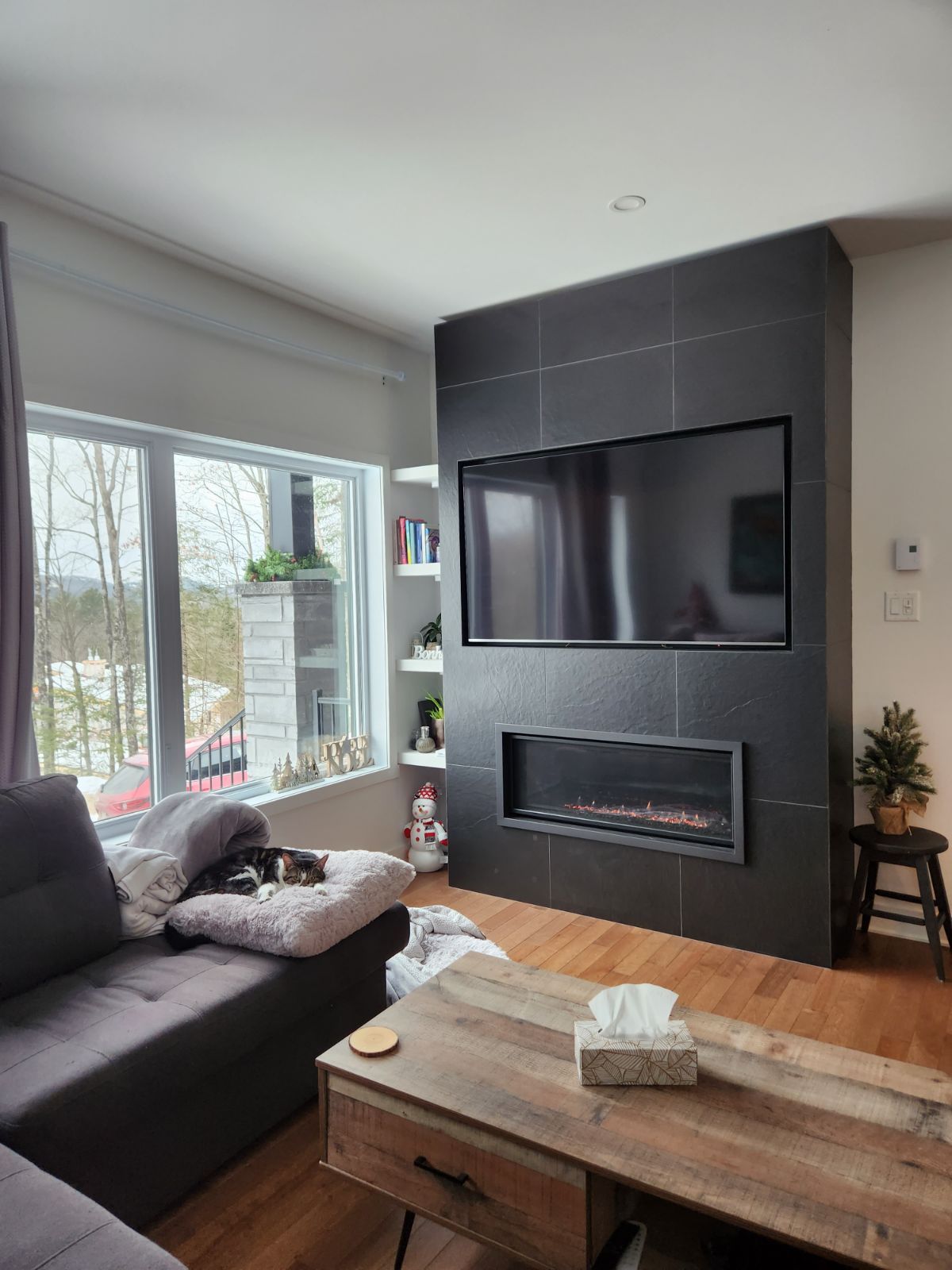 Salon avec cheminée, télévision et canapé d'angle près d'une fenêtre donnant sur les arbres.