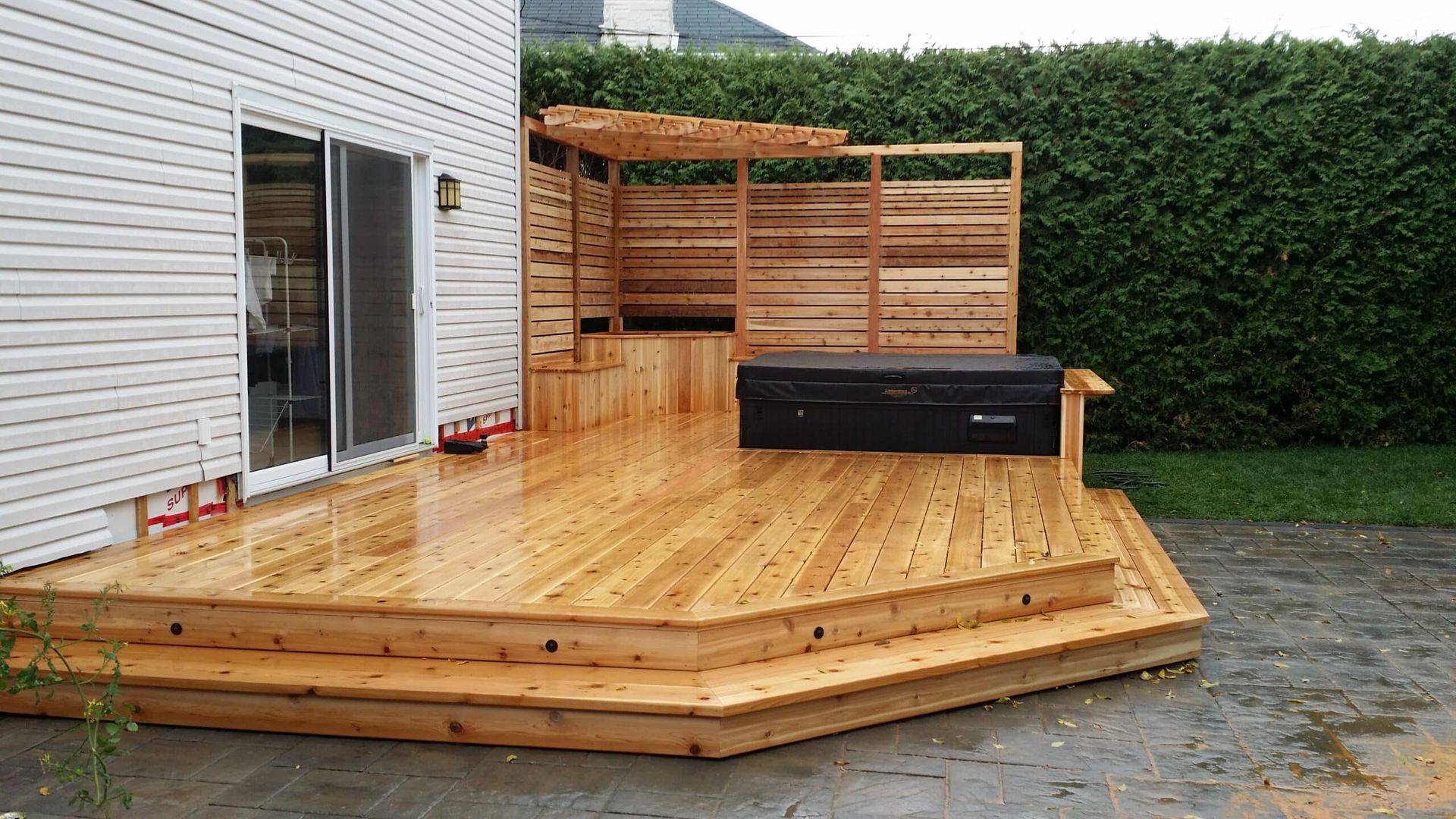 Terrasse en bois avec jacuzzi intégré, pergola et escalier, jouxtant une maison avec portes coulissantes en verre.