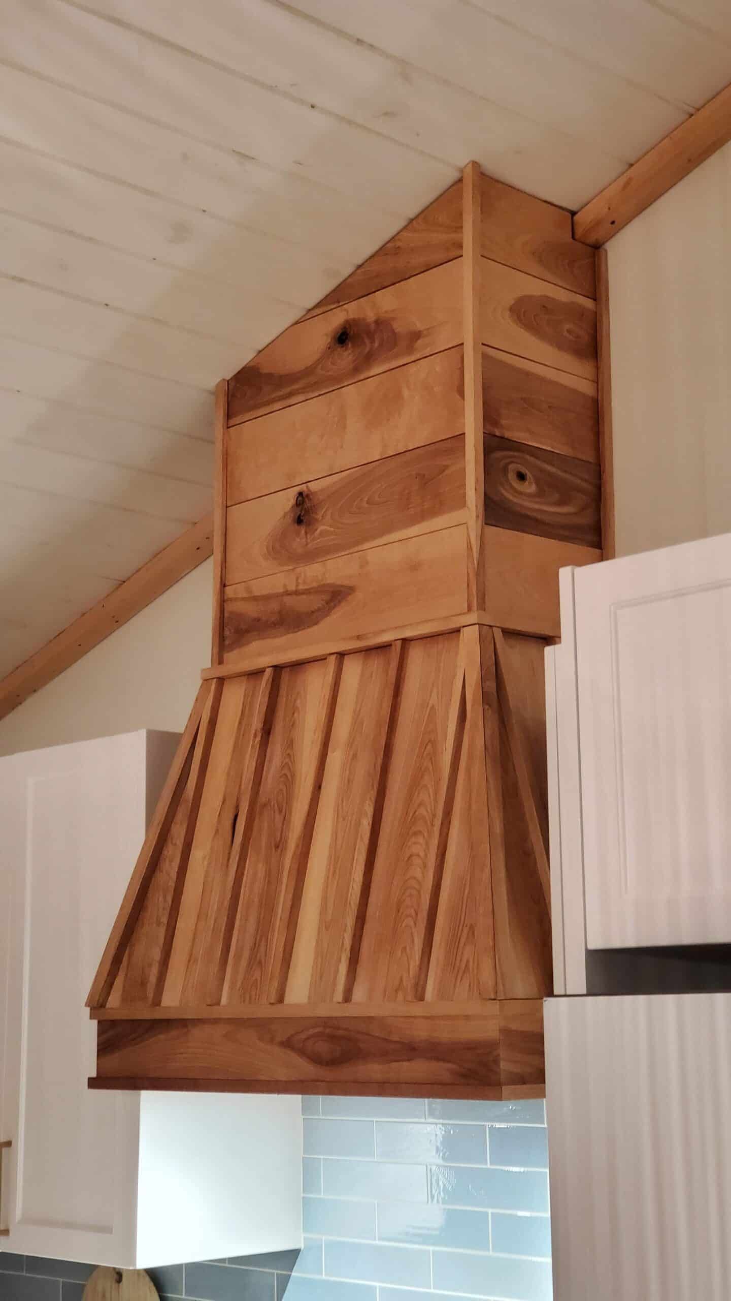 Hotte de cuisine en bois à façade inclinée, montée entre des armoires blanches et contre un plafond clair.