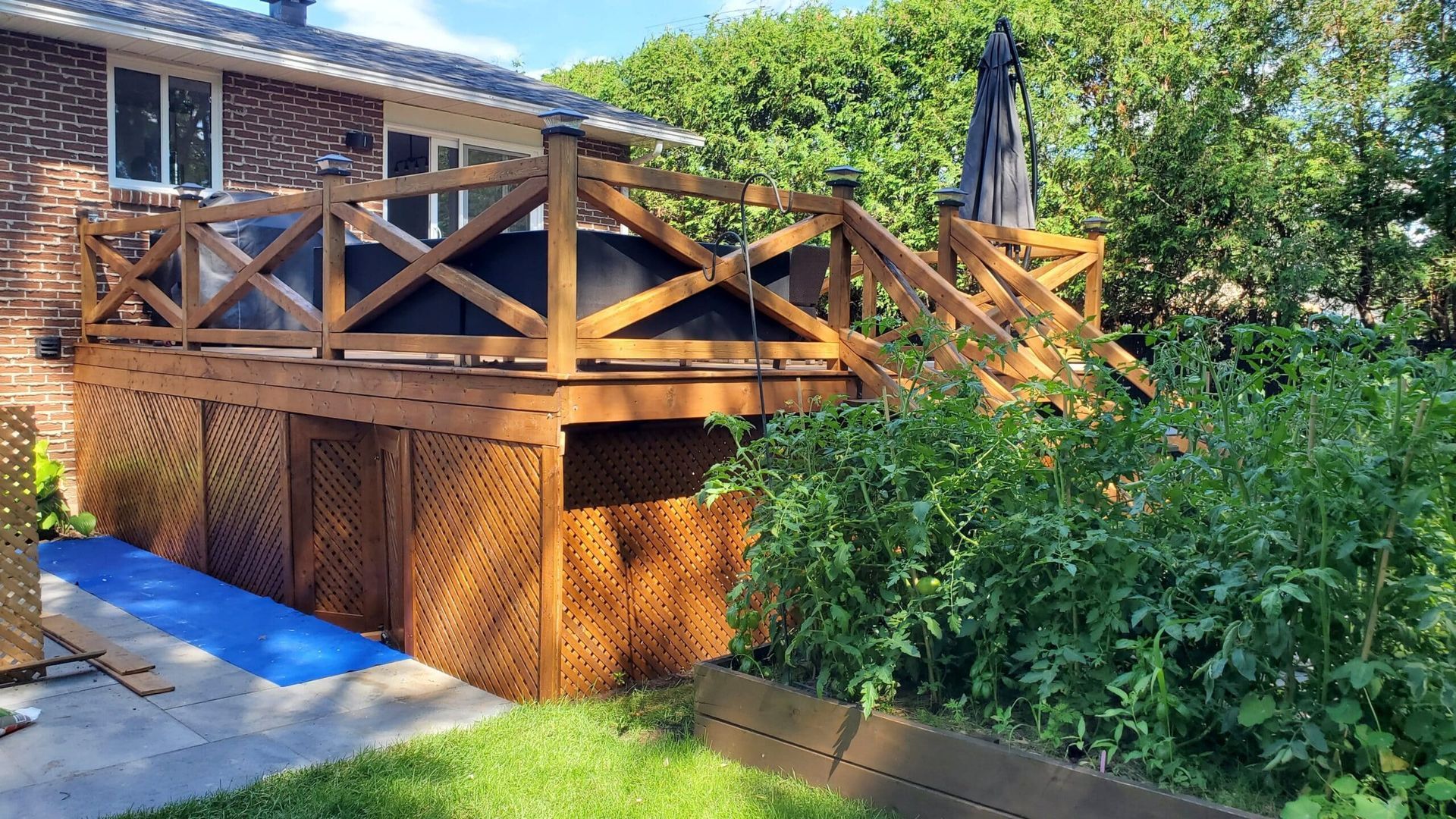 Terrasse en bois avec treillis et rambarde, attenante à une maison en briques. La terrasse est entourée de végétation.