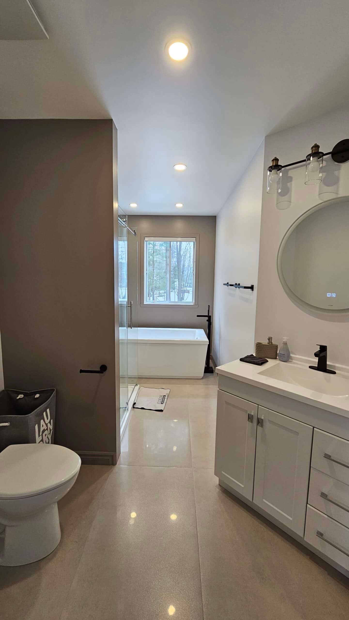 Salle de bains moderne avec meuble-lavabo, toilettes et baignoire blancs ; murs gris et sol clair.