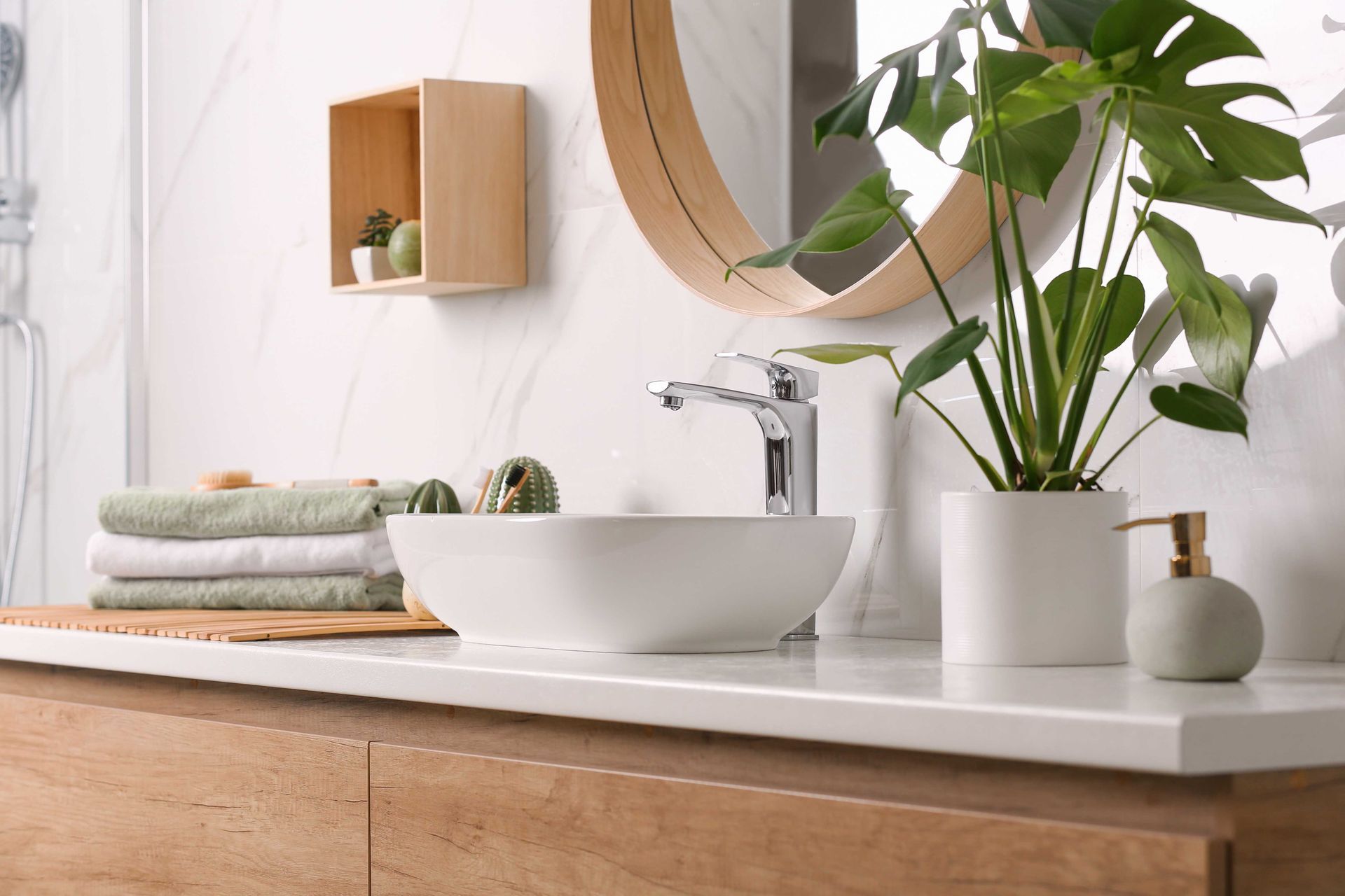 Salle de bains avec comptoir blanc, meuble en bois, miroir rond et plante en pot.