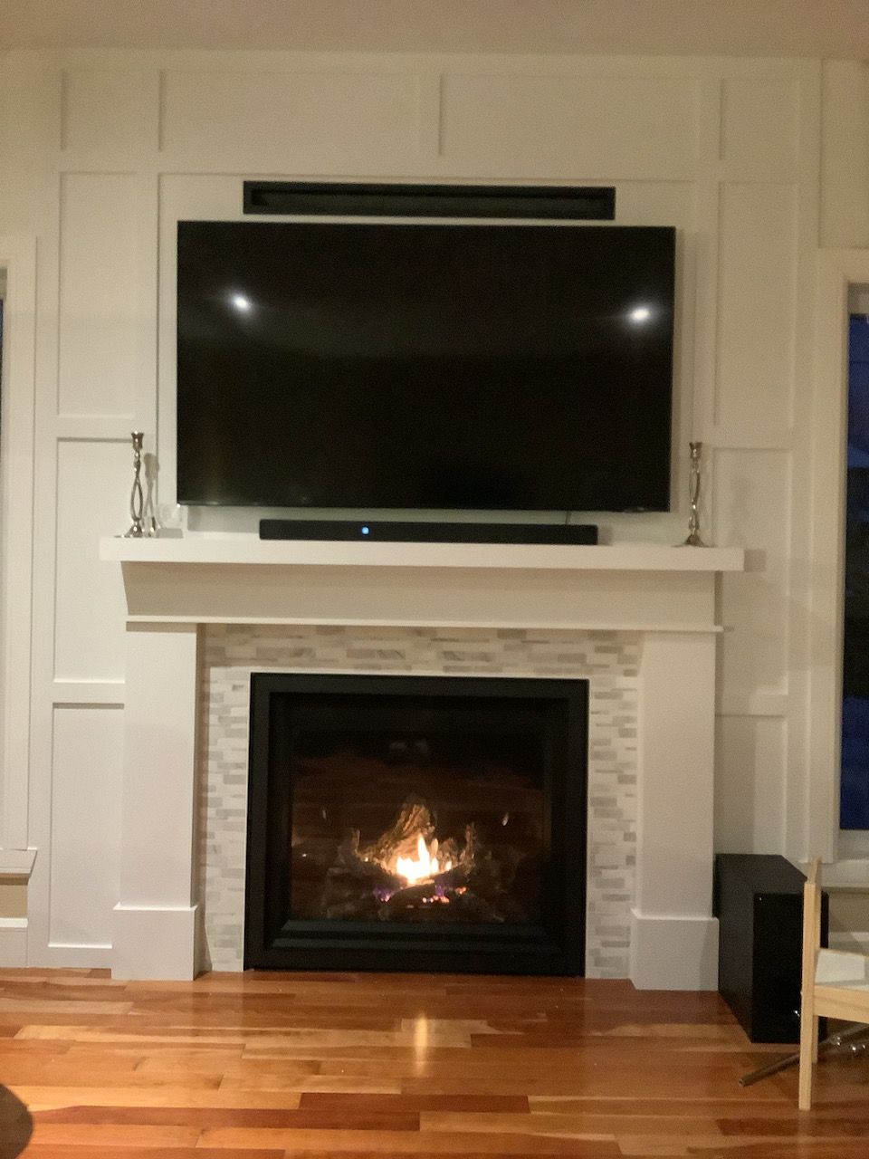 Cheminée avec feu allumé, télévision et barres de son, dans un décor lambrissé blanc, avec parquet.