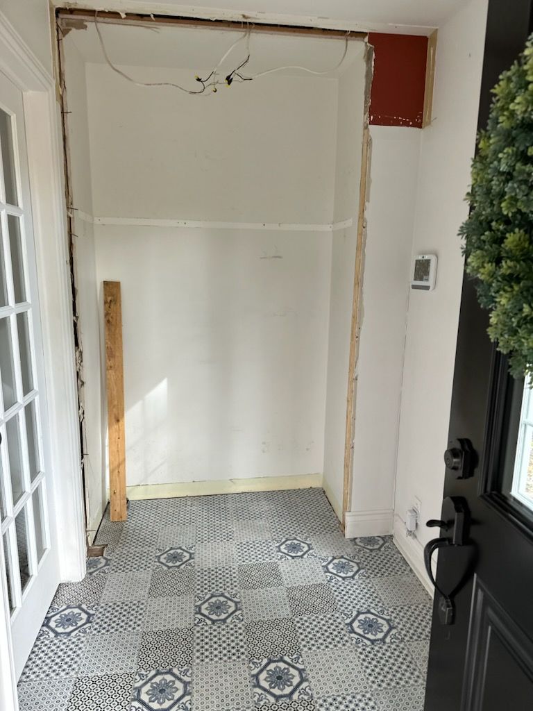 Rénovation de l'entrée. Montants muraux apparents, carrelage à motifs, murs blancs, câblage visible, porte partiellement fermée et couronne.