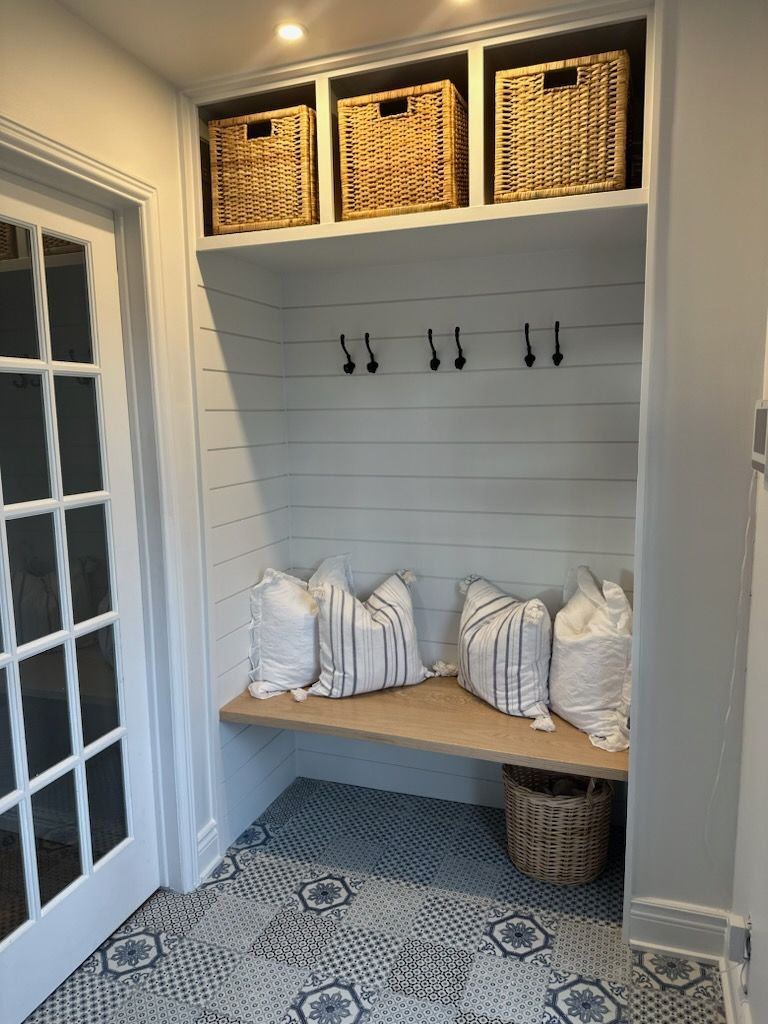 Vestibule intégré avec banc, patères, paniers tressés et sol à motifs. Mur en lambris blanc et trois coussins.
