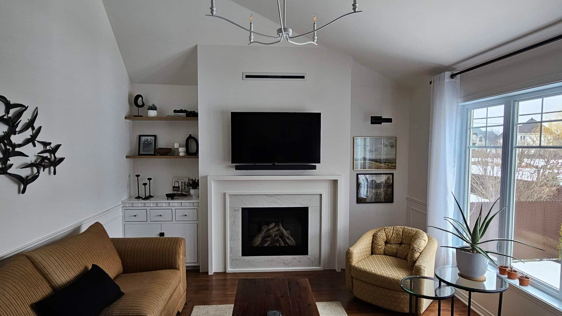 Salon avec cheminée, télévision, canapé, fauteuil et étagères, avec une fenêtre et un lustre moderne.