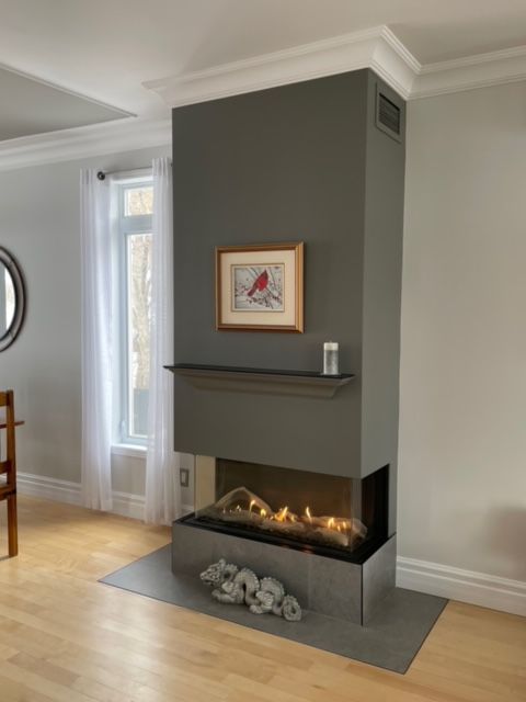 Cheminée grise moderne avec œuvres d'art, étagère et flammes de gaz dans une pièce d'angle avec fenêtre et parquet.