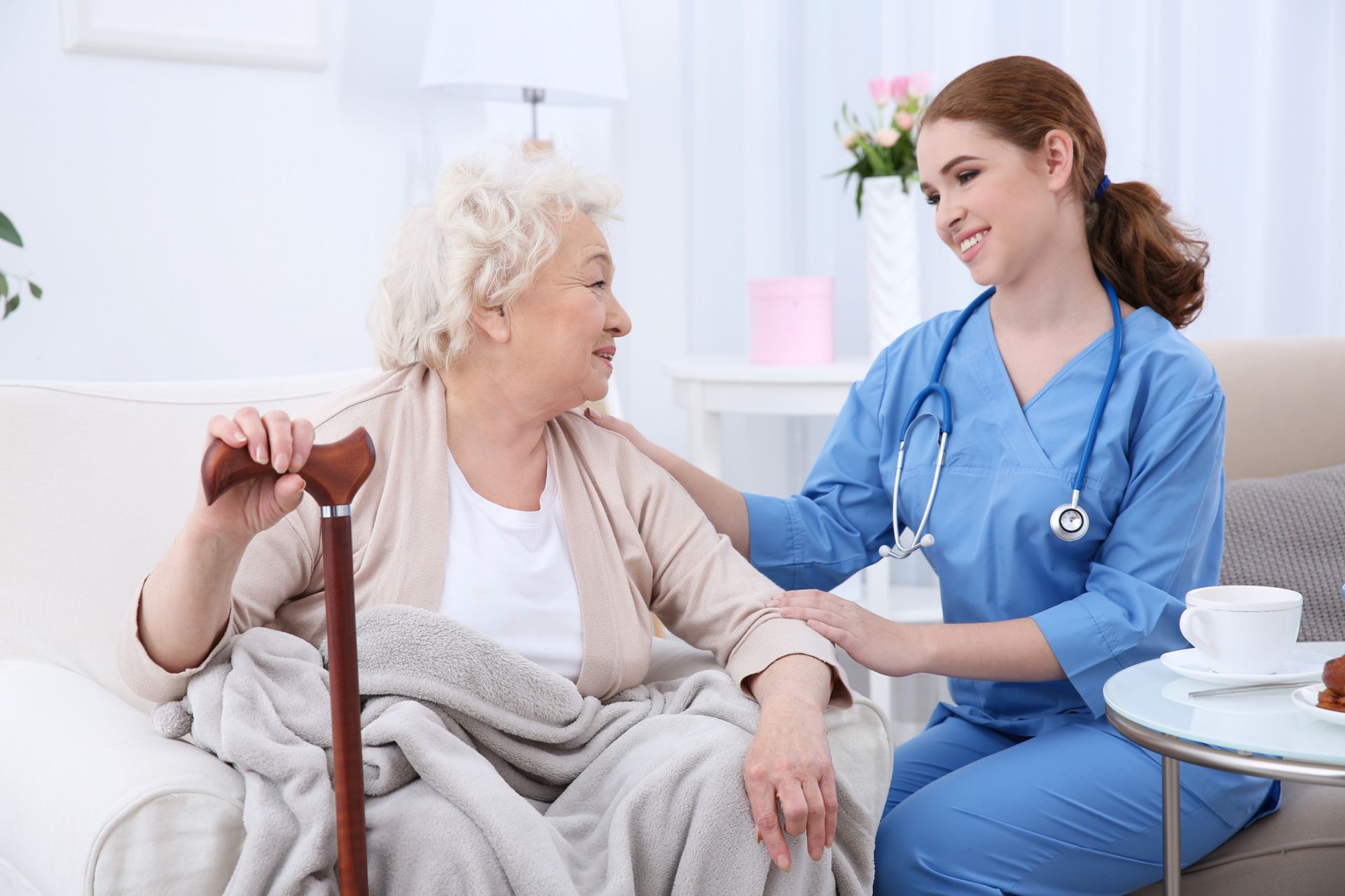 A caregiver in blue scrubs smiles while comforting an elderly person sitting with a cane and blanket in a home setting.