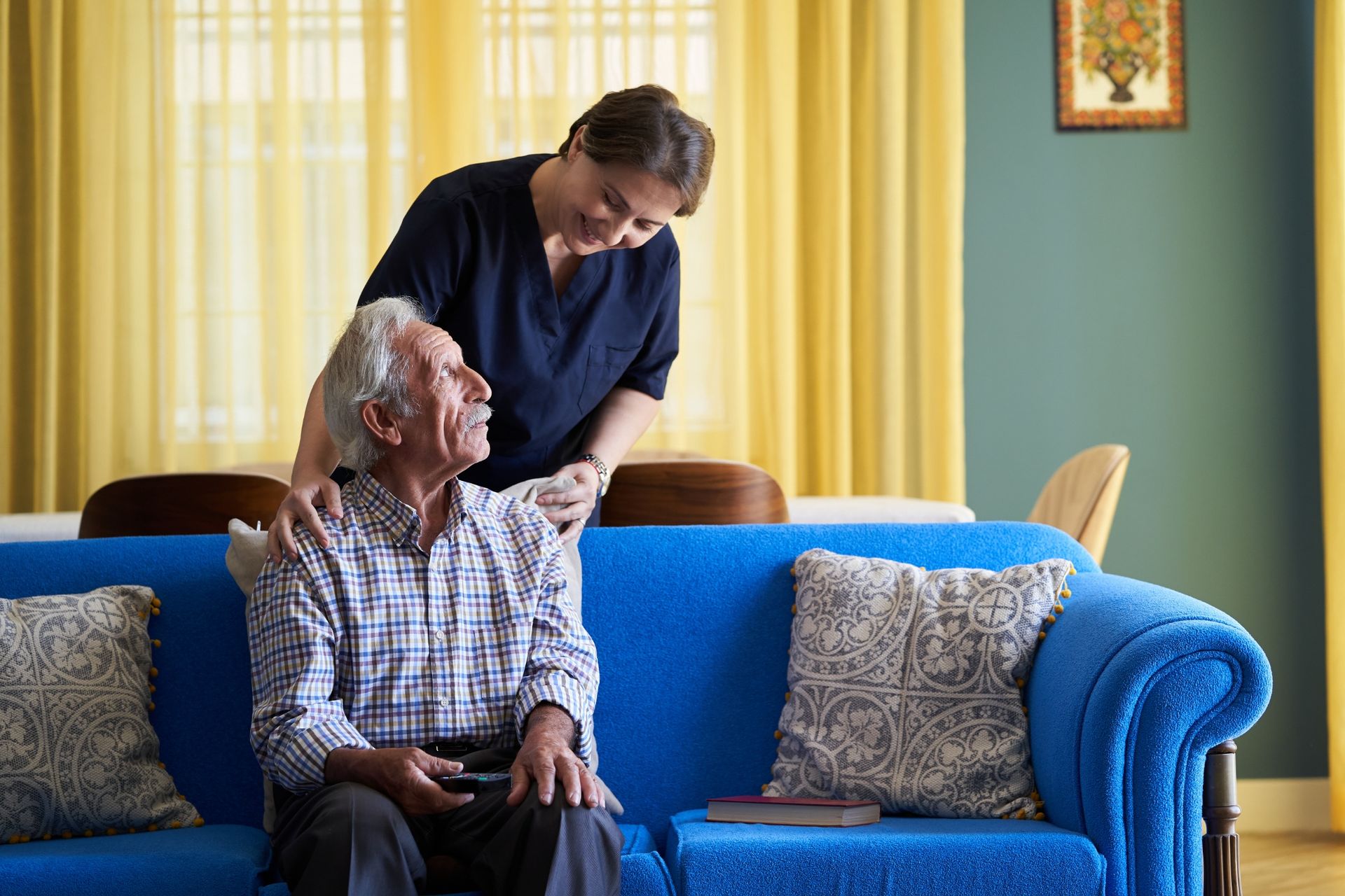 A person in dark blue scrubs stands behind an elderly person seated on a bright blue sofa, resting hands on their shoulders.