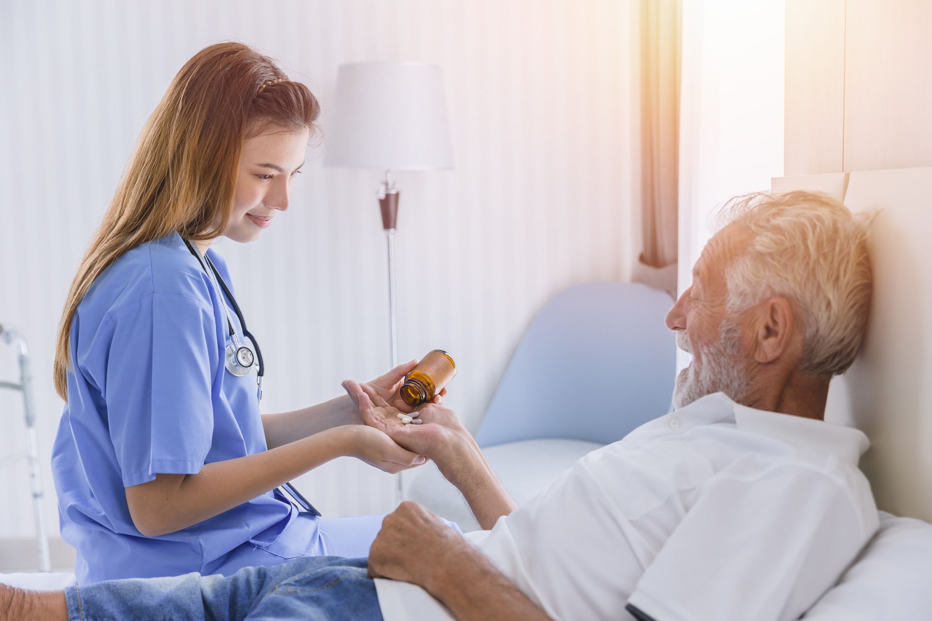 A healthcare provider in blue scrubs hands medication to an older person resting in a bed.
