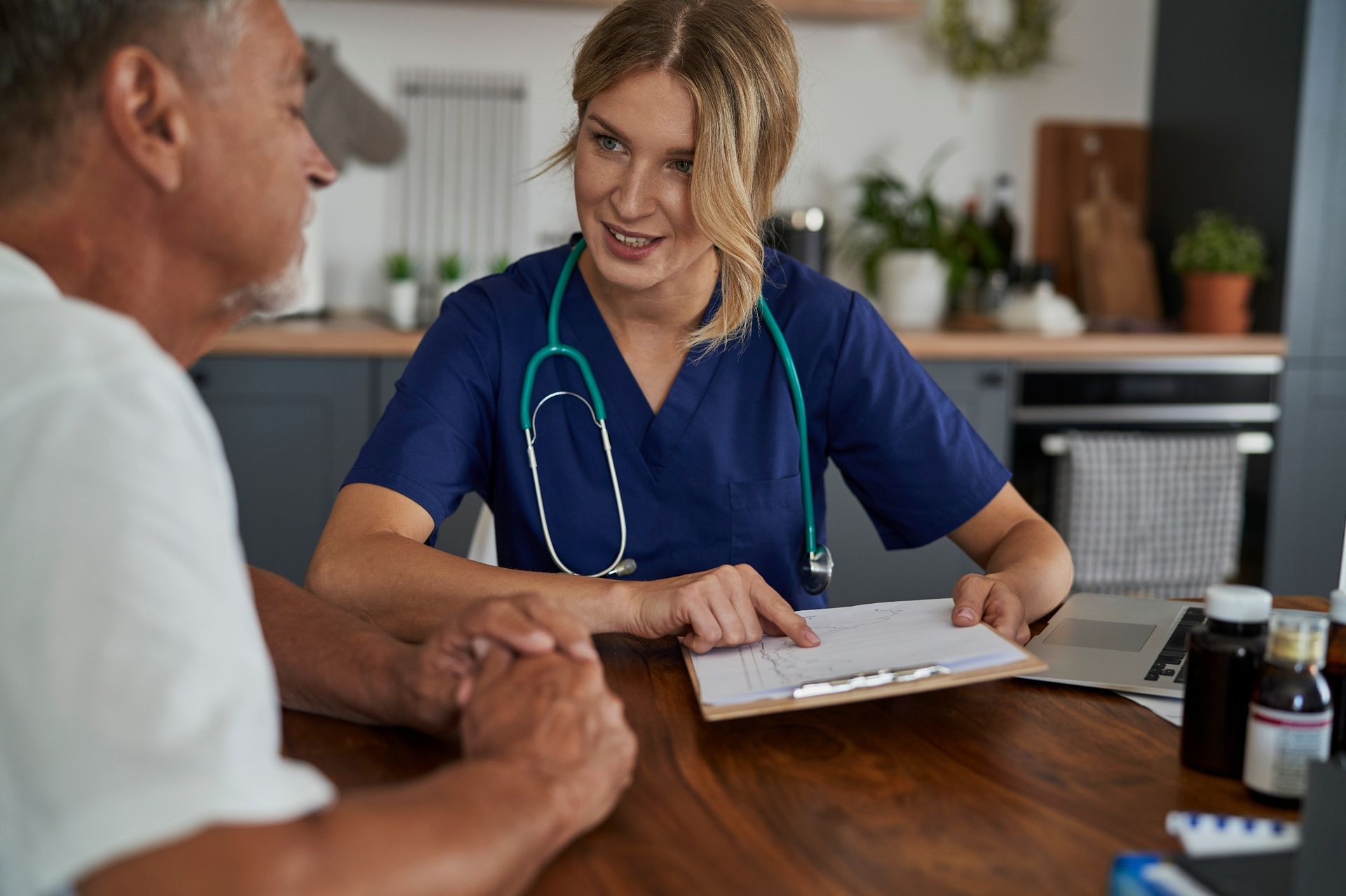A healthcare professional in scrubs reviews paperwork with a patient at a wooden table in a home setting.