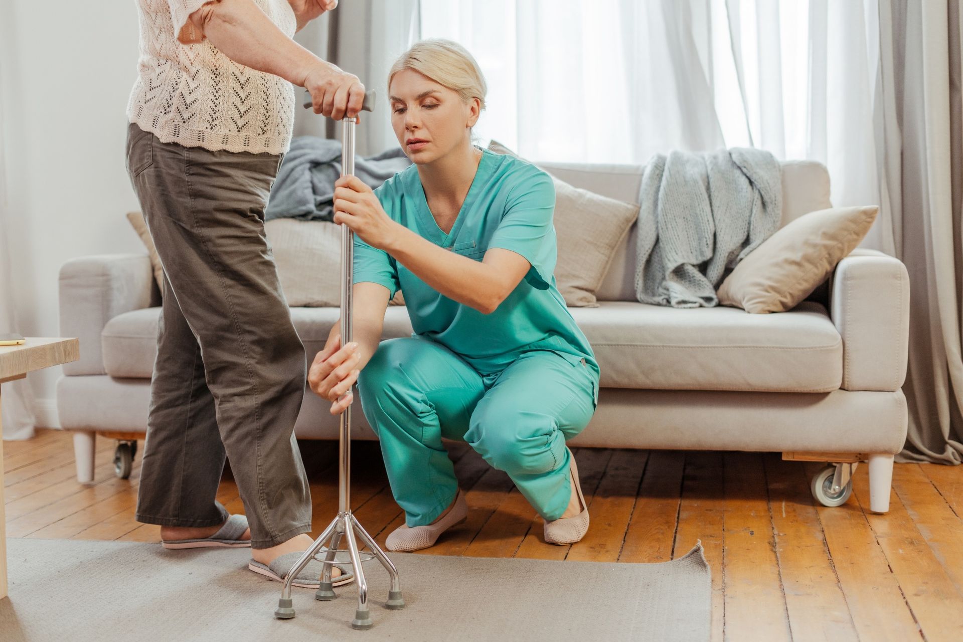 A healthcare worker in teal scrubs assists an individual with standing by using a four-pronged metal cane in a home room.