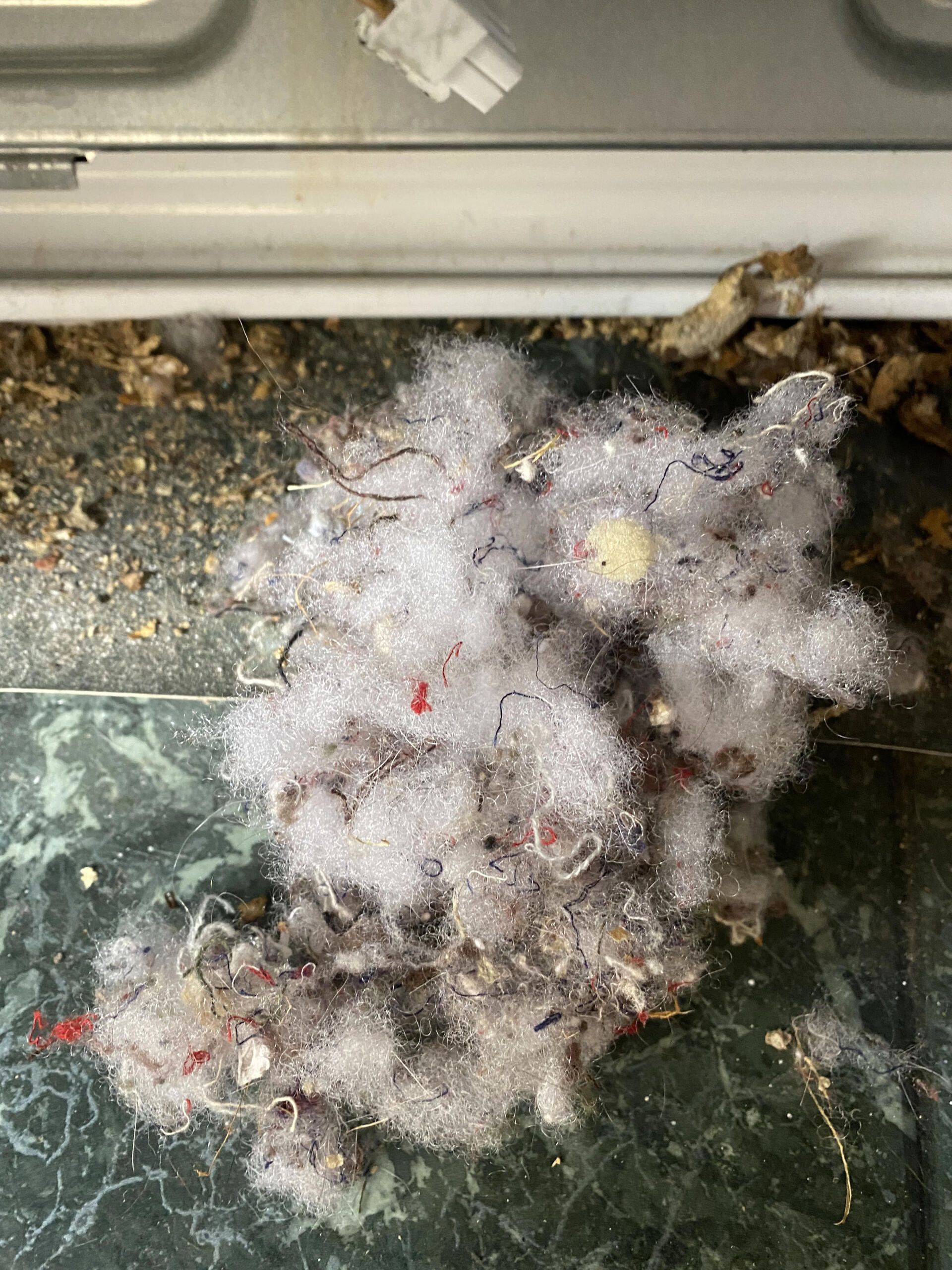 A pile of gray dryer lint sits on a textured surface in front of a white appliance panel.