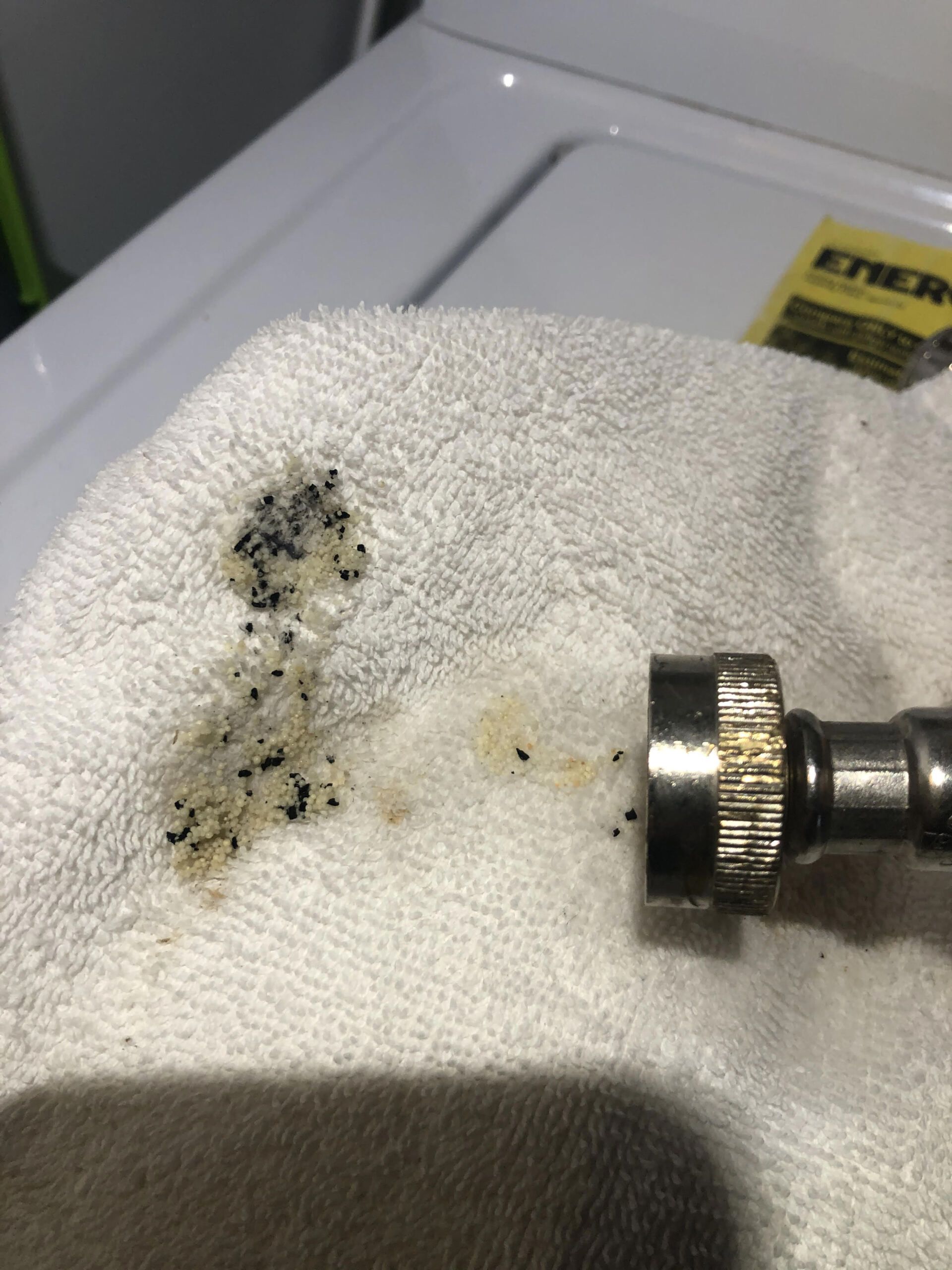 A metal water hose connector resting on a white towel, with black and tan sediment debris spilled onto the fabric.