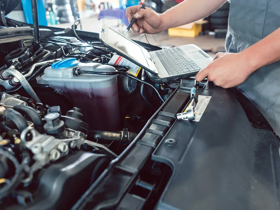 A mechanic uses a laptop and diagnostic tool to inspect an engine bay in an automotive shop.