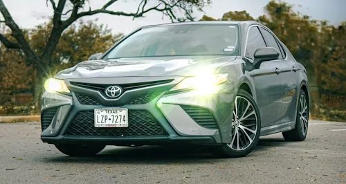 Gray Toyota Camry sedan with headlights on, parked outdoors. Texas license plate visible.