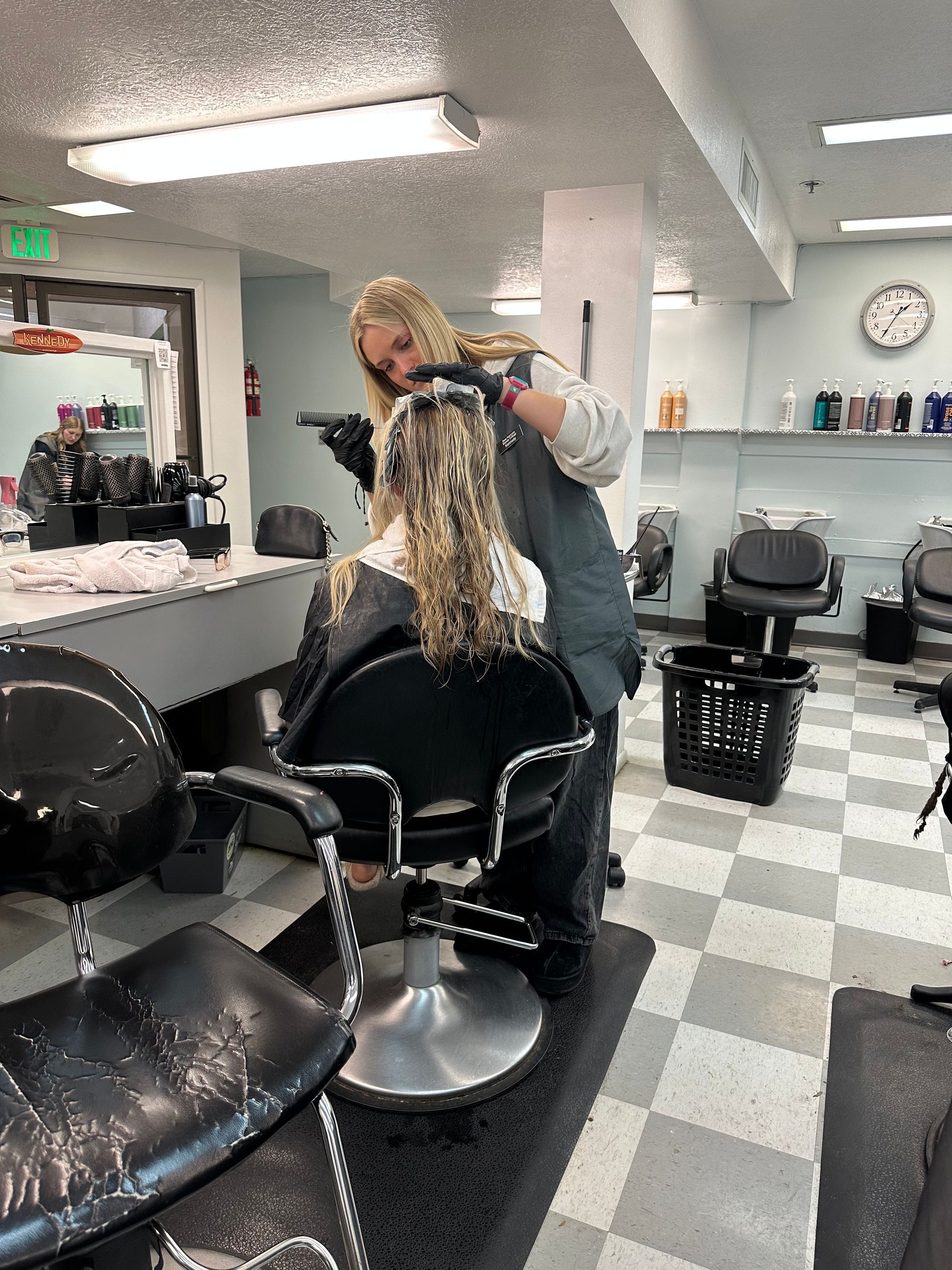 A stylist in a salon applying treatment to a client's blonde, curly hair, seated in a chair on a checkered floor. A stylist in a salon applying treatment to a client's blonde, curly hair, seated in a chair on a checkered floor.