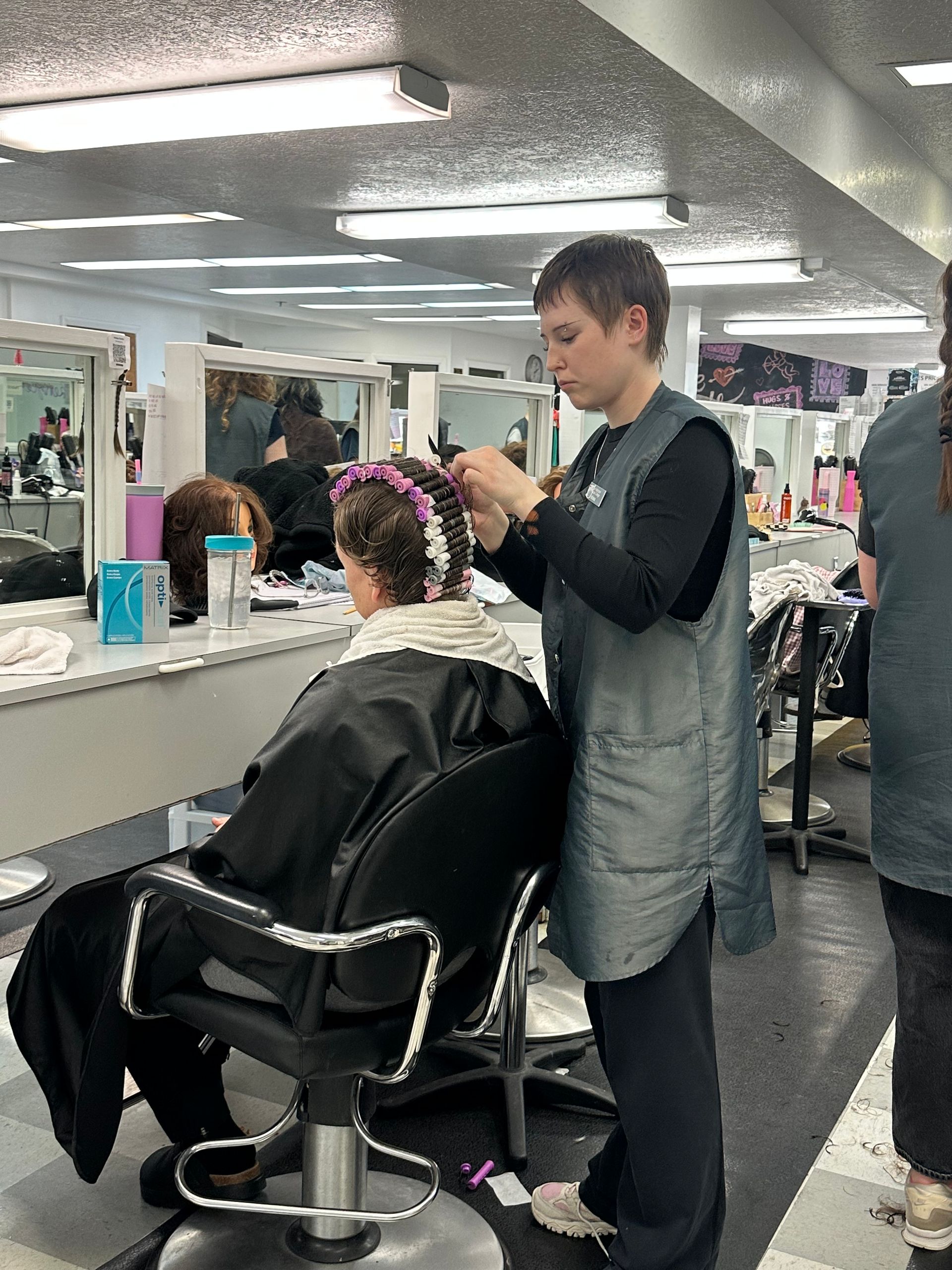 A person in a gray vest styles another person's hair with rollers in a brightly lit hair salon.