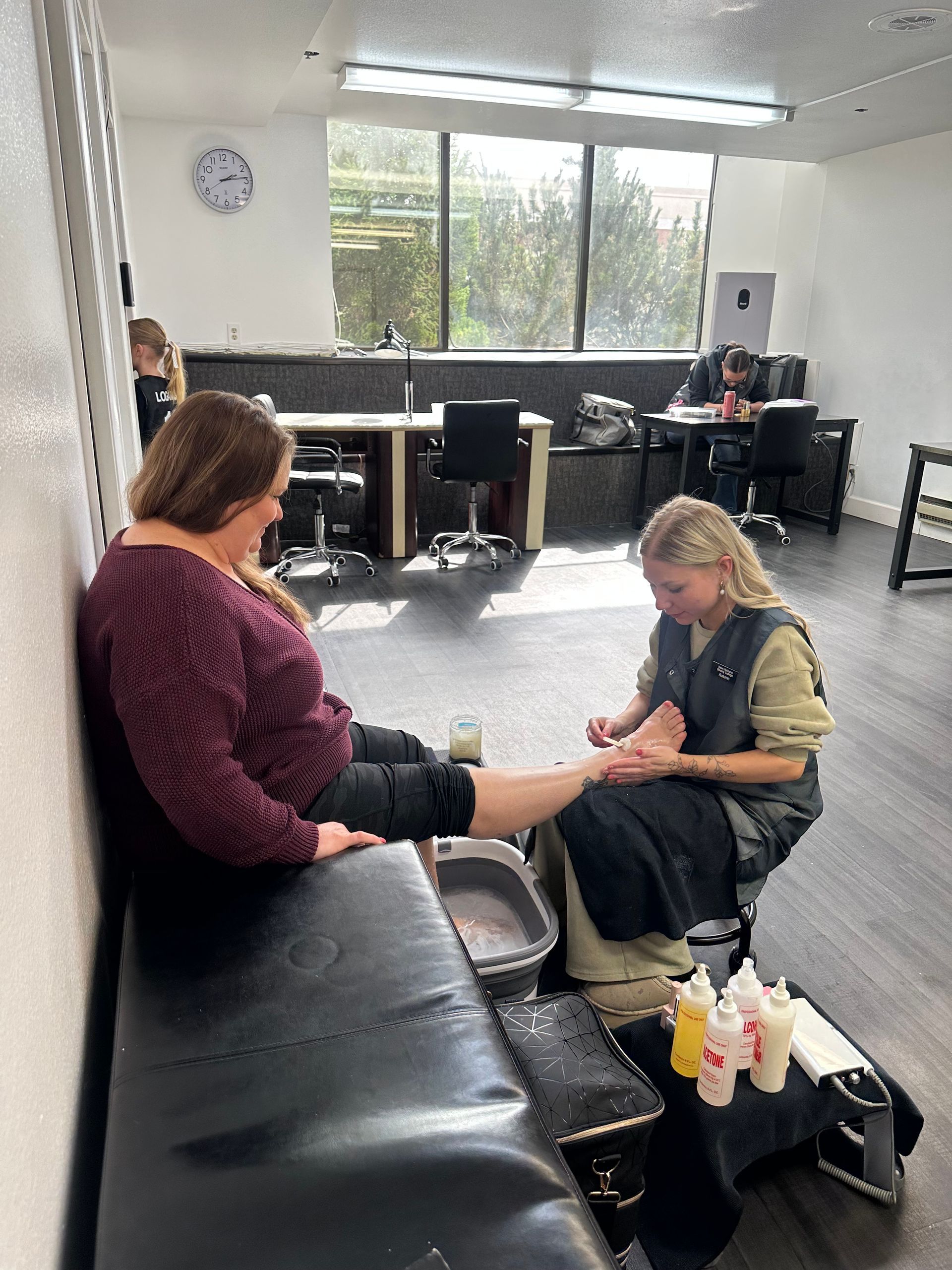 A person in a maroon sweater receives a pedicure from a technician in a grey vest inside a brightly lit salon.